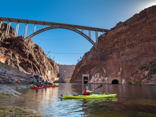 A kayaker paddles through the calm waters of Black Canyon near Hoover Dam, with the impressive Mike O'Callaghan–Pat Tillman Memorial Bridge towering overhead.
