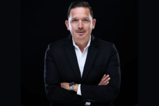 A man in a dark suit with a white shirt, arms crossed, against a black background. He has a confident expression, conveying professionalism.