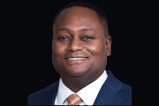 Professional headshot of a smiling Black man in a navy suit, white shirt, and patterned tie against a black background, conveying warmth and confidence.